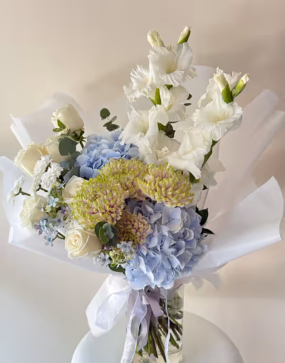 Bouquet with white roses, white gladiolus, light green sedum, blue hydrangeas, and small white and blue flowers in a clear glass vase with white wrapping.