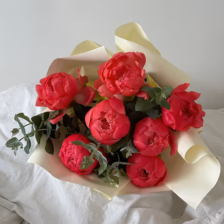 Bouquet of red peony flowers wrapped in pale yellow paper resting on white fabric.