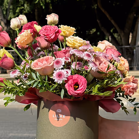 Bouquet of mixed pink, yellow, and white flowers arranged with greenery in a green basket with a red ribbon and Lilae label.