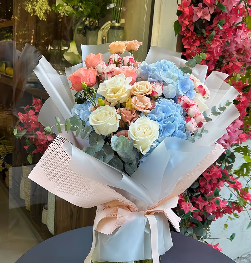 Bouquet of white roses, blue hydrangeas, and pink flowers wrapped in translucent paper with ribbons.