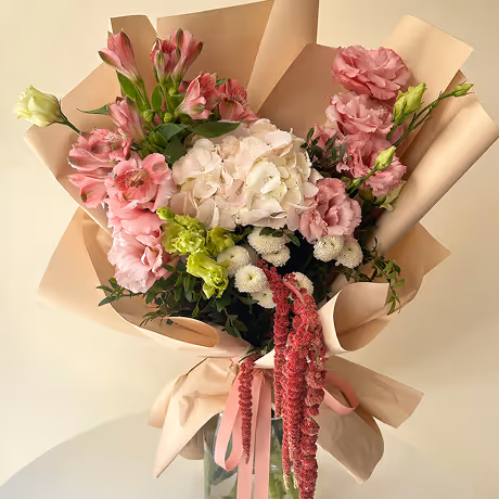Bouquet of pink and white flowers including hydrangeas and carnations wrapped in beige paper with a pink ribbon.