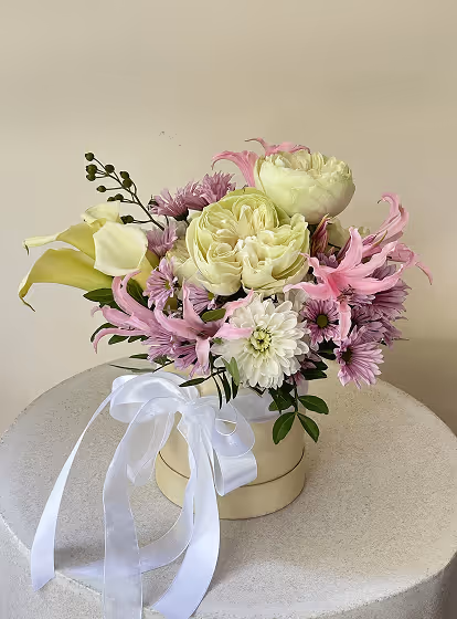 Bouquet of pale yellow roses, white chrysanthemums, pink lilies, and purple flowers arranged in a round cream container with a white ribbon on a round table.