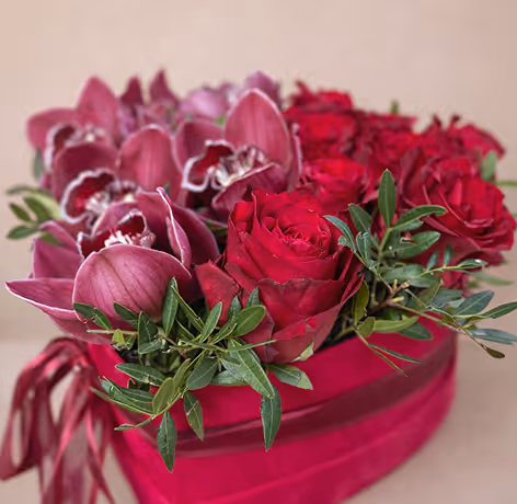 Bouquet of red roses and purple orchids with green leaves in a red heart-shaped box.