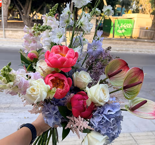 Hand holding a colorful bouquet with pink peonies, white roses, purple hydrangeas, and green foliage outdoors.