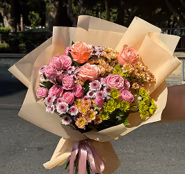 Hand holding a bouquet of pink roses, purple chrysanthemums, and green foliage wrapped in beige paper with a pink ribbon.