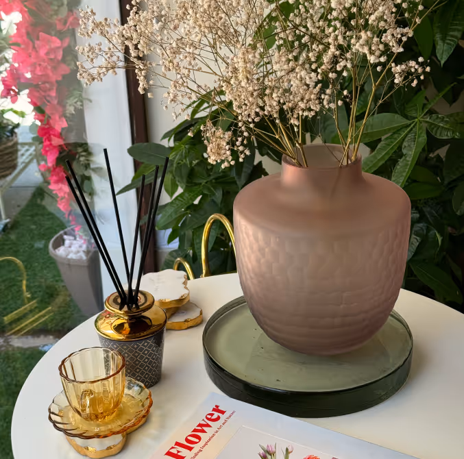 Decorative pinkish vase with dried baby's breath flowers on a glass plate next to a reed diffuser and amber glass teacup on a white round table.
