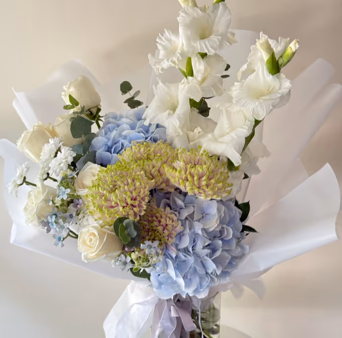 Bouquet with white roses, white gladiolus, blue hydrangeas, and greenish-yellow textured flowers wrapped in white paper.