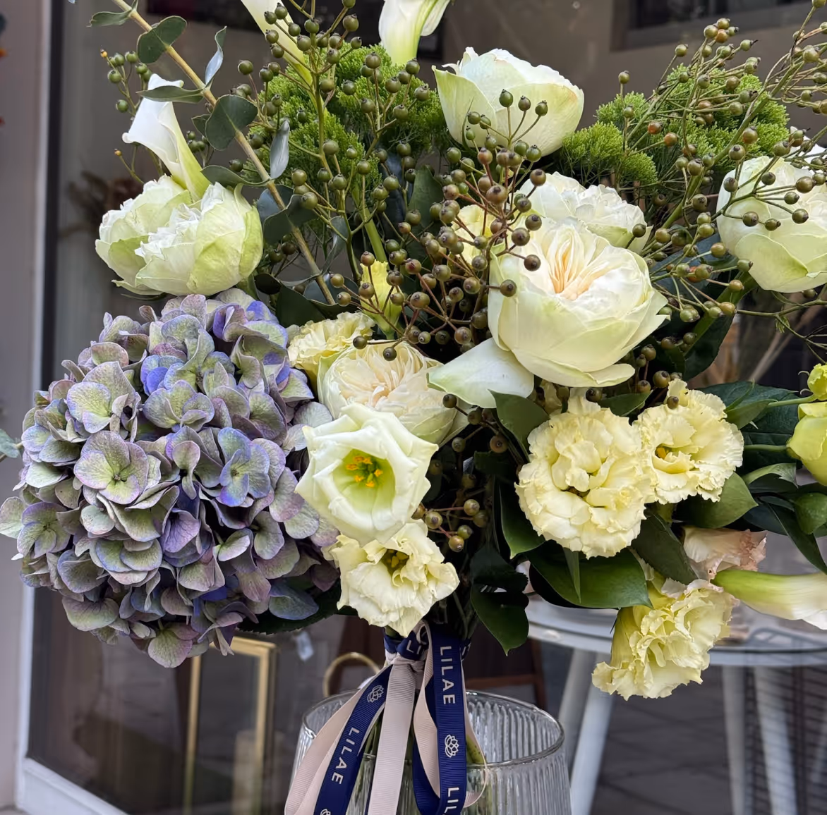 Bouquet of cream roses, pale yellow carnations, green foliage, and a cluster of purple hydrangeas.