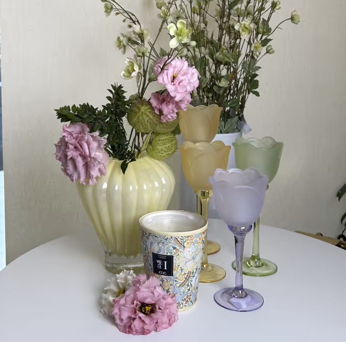 Arrangement of pink and white flowers in a pale yellow vase and a blue floral-patterned cup on a white table with three pastel-colored frosted glass goblets in the background.