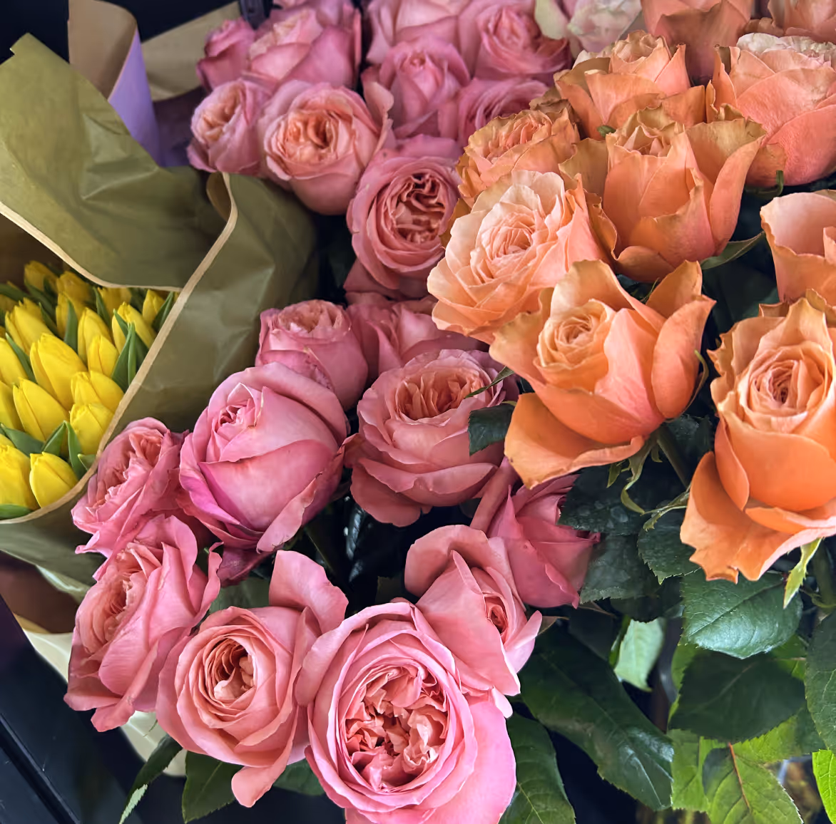Close-up of bouquets with pink and peach roses, and yellow tulips wrapped in green paper.
