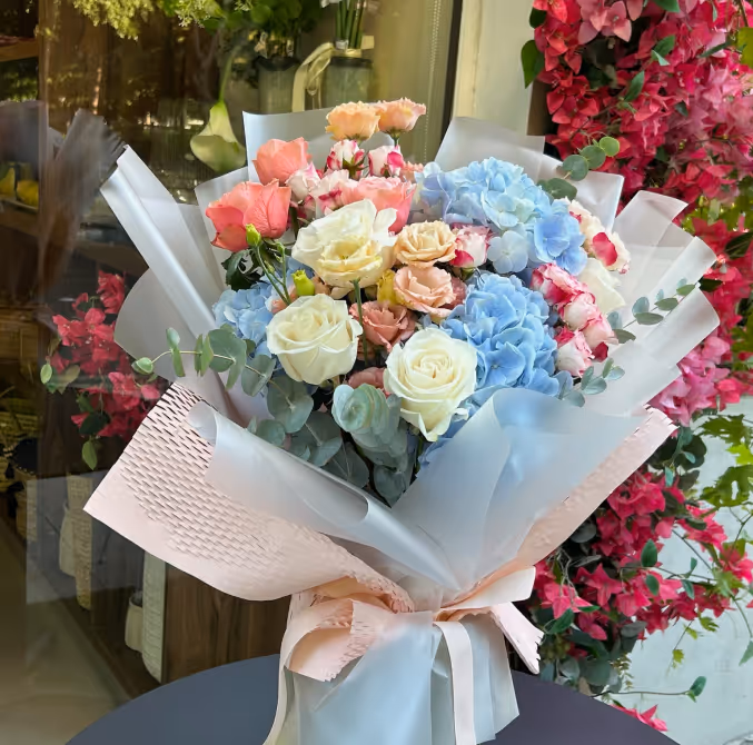 Bouquet of mixed pastel flowers including white roses, blue hydrangeas, and pink spray roses wrapped in translucent white and light pink paper with eucalyptus leaves.