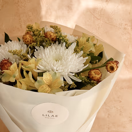 Bouquet of white chrysanthemums, yellow lilies, and dried flowers wrapped in beige paper with a Lilae label.