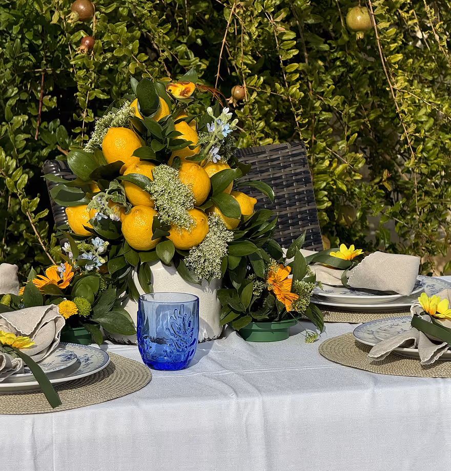 Table set with plates, napkins, a blue glass, and a centerpiece of lemons and green leaves outdoors.