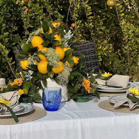 Outdoor dining table set with beige napkins and yellow flowers, featuring a centerpiece of fresh lemons and greenery in a white vase.