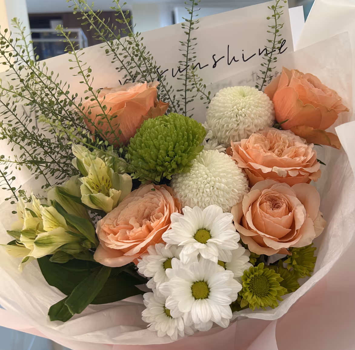 Bouquet of peach roses, white daisies, green pom-poms, and assorted greenery wrapped in white paper.