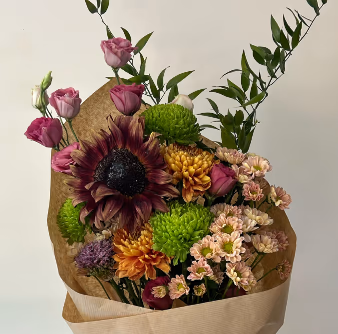 Bouquet of mixed flowers including purple sunflower, orange chrysanthemums, green pom-poms, pink roses, and leafy greens wrapped in brown paper.
