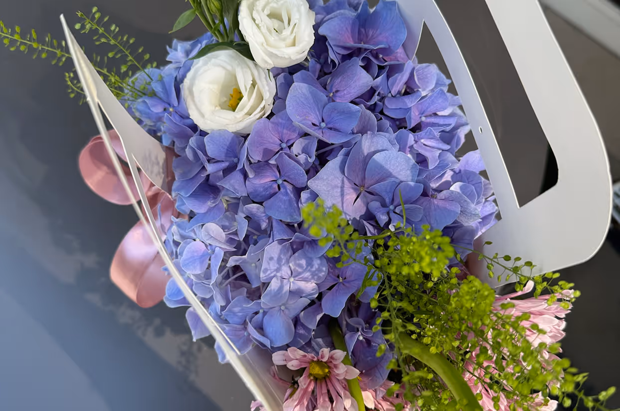 Bouquet with purple hydrangeas, white roses, pink daisies, and green foliage in a white holder.