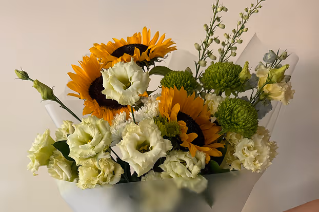 Bouquet featuring bright yellow sunflowers, white lisianthus, green pom-pom chrysanthemums, and assorted greenery wrapped in translucent paper.
