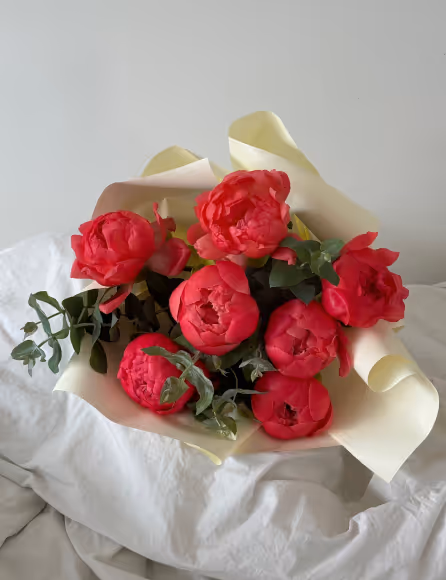 Bouquet of bright red peony flowers wrapped in cream paper on a white fabric surface.