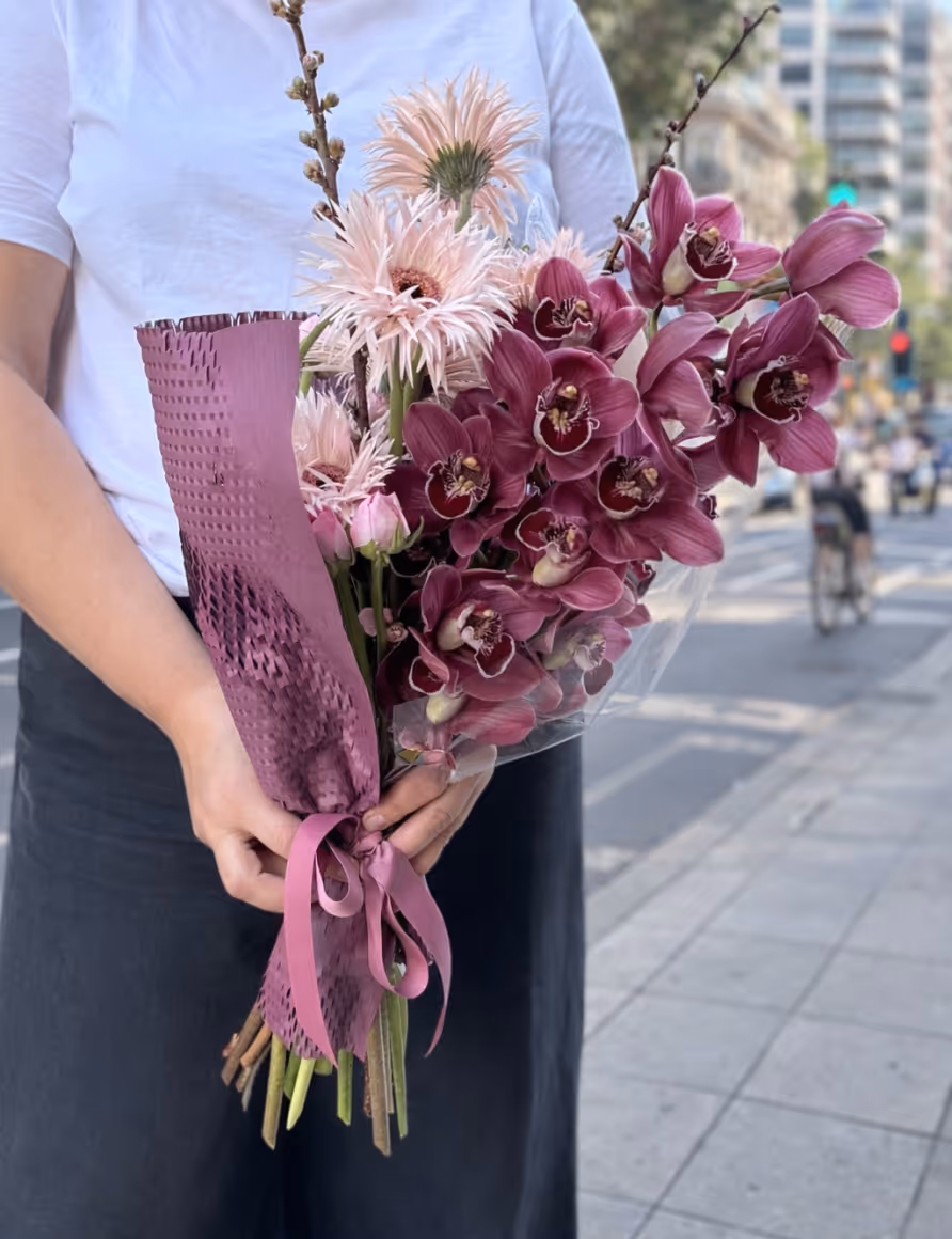 Person holding a bouquet of pink and purple flowers wrapped in textured purple paper with a ribbon.