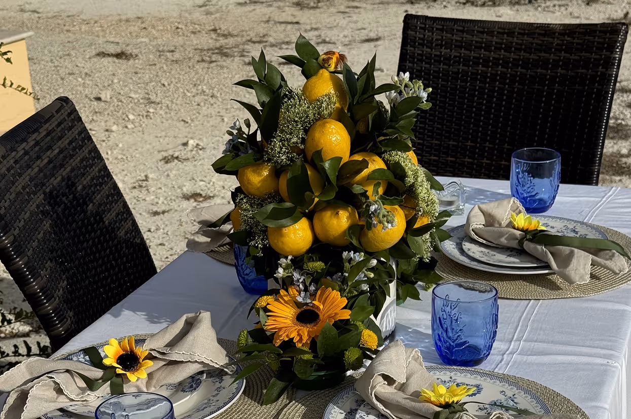 Table set outdoors with lemon-themed centerpiece, blue glasses, sunflowers, and napkins on floral plates.