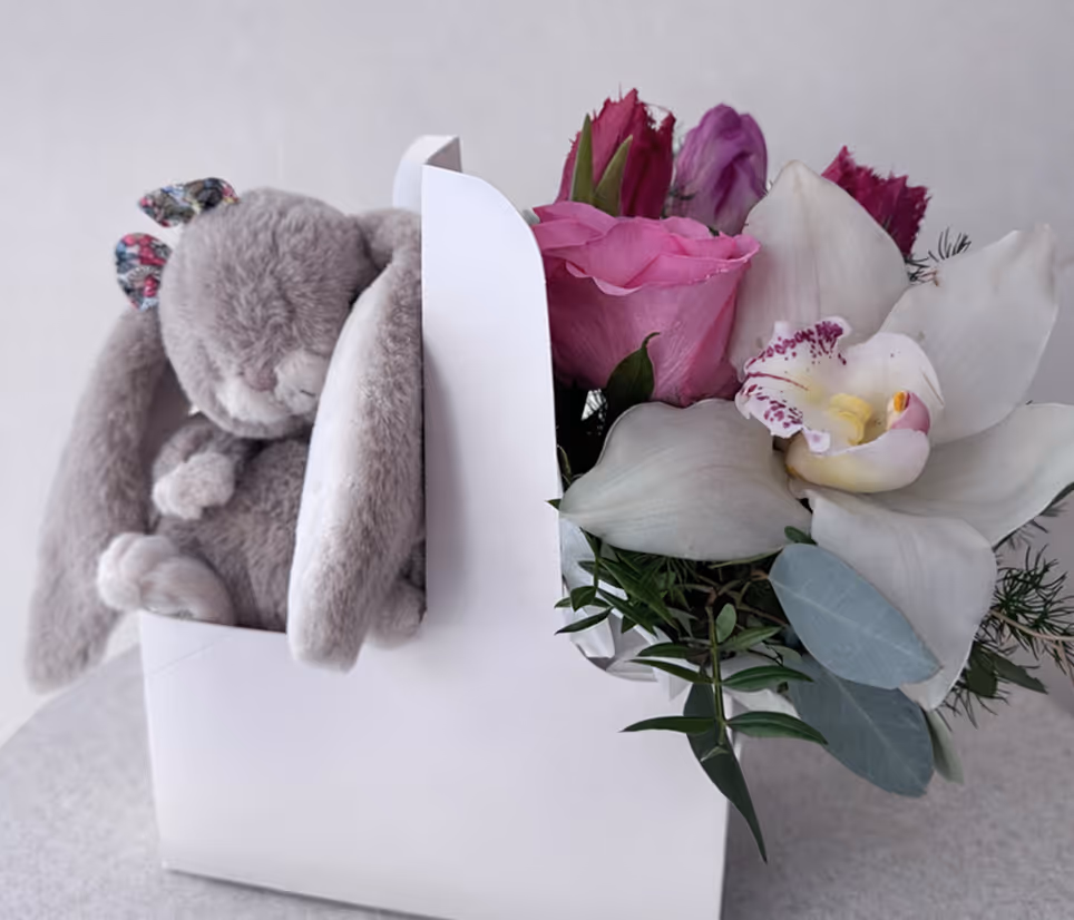 Gray plush bunny with floppy ears next to pink and white flowers in a white box.