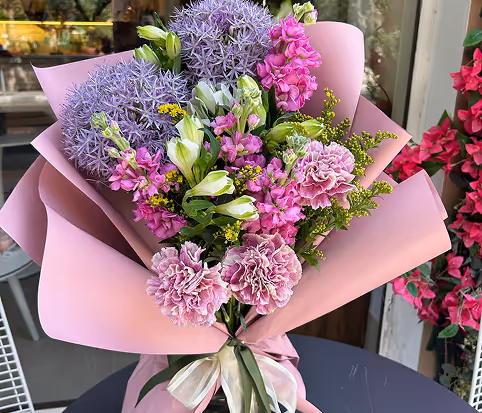 Bouquet with purple allium, pink carnations, small pink flowers, and green foliage wrapped in pink paper with a white ribbon.