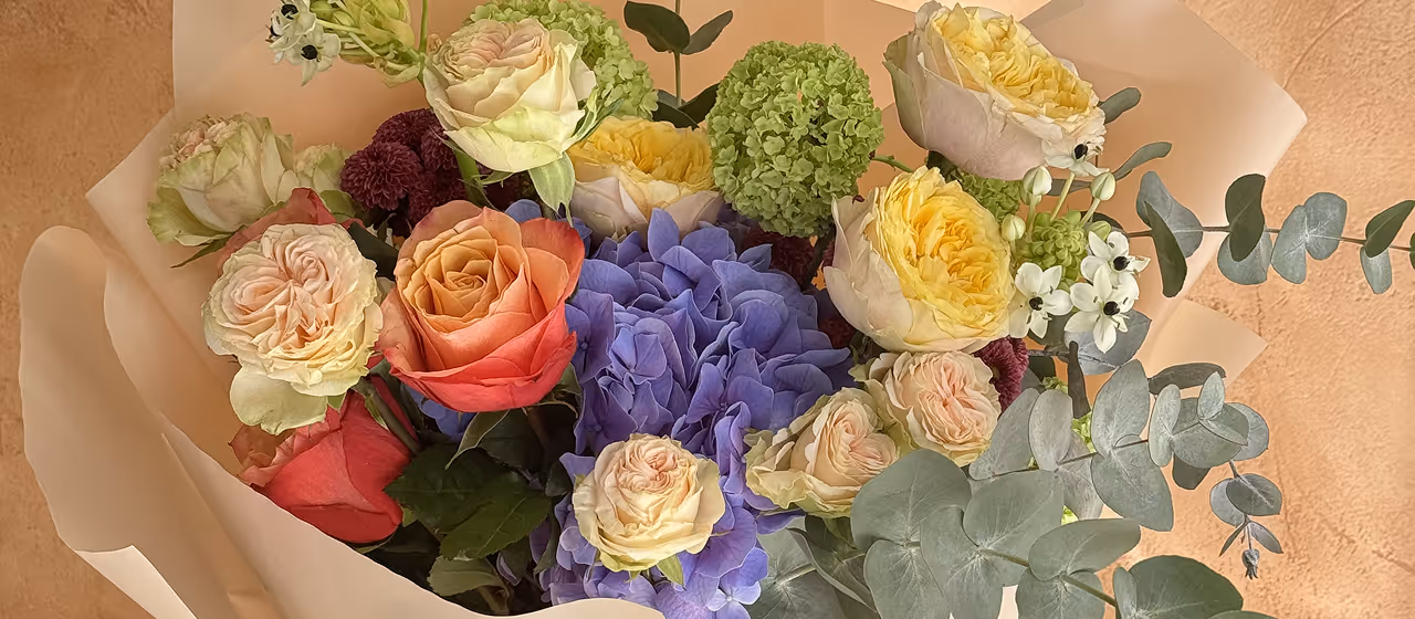 Bouquet featuring cream and peach roses, purple hydrangeas, green viburnum, white blossoms, and eucalyptus leaves wrapped in beige paper.