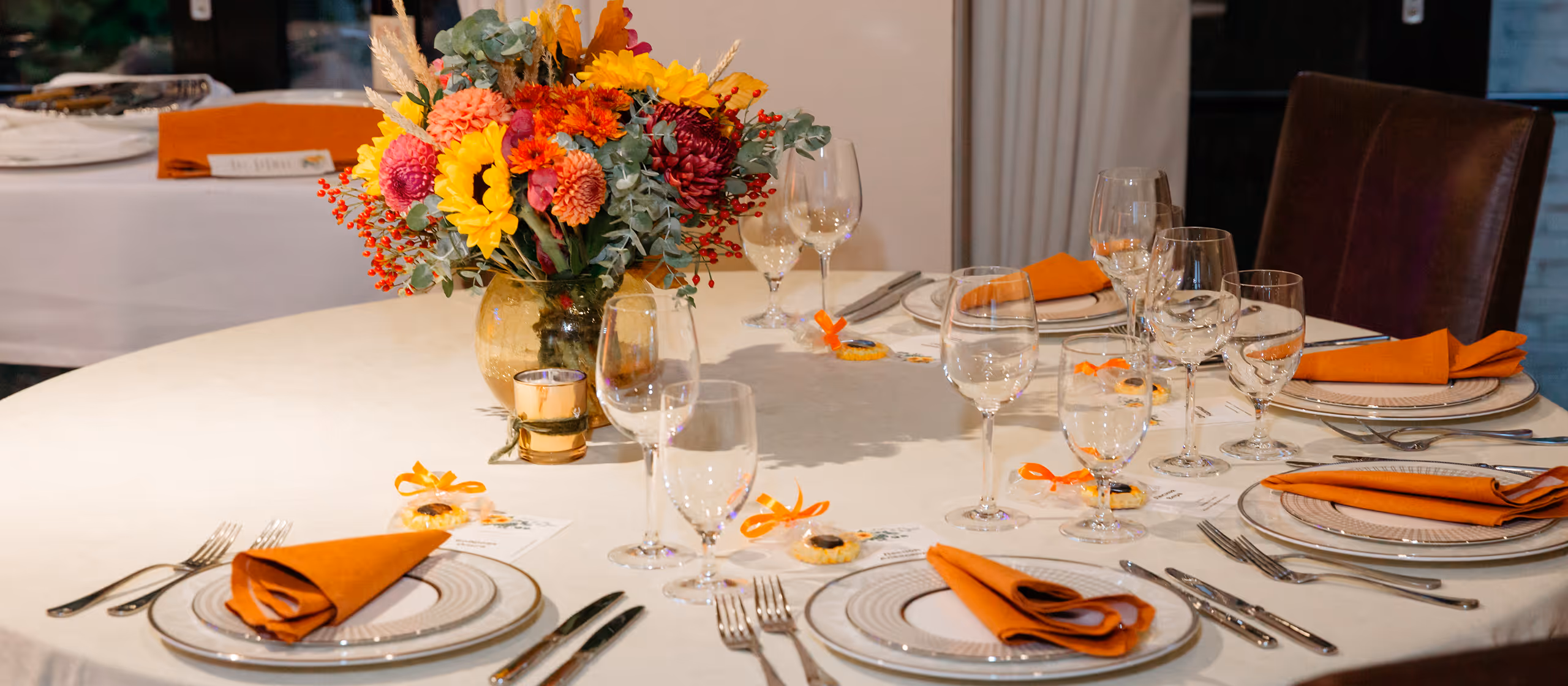 Elegant round dining table with white tablecloth, floral centerpiece, orange napkins, and glassware.