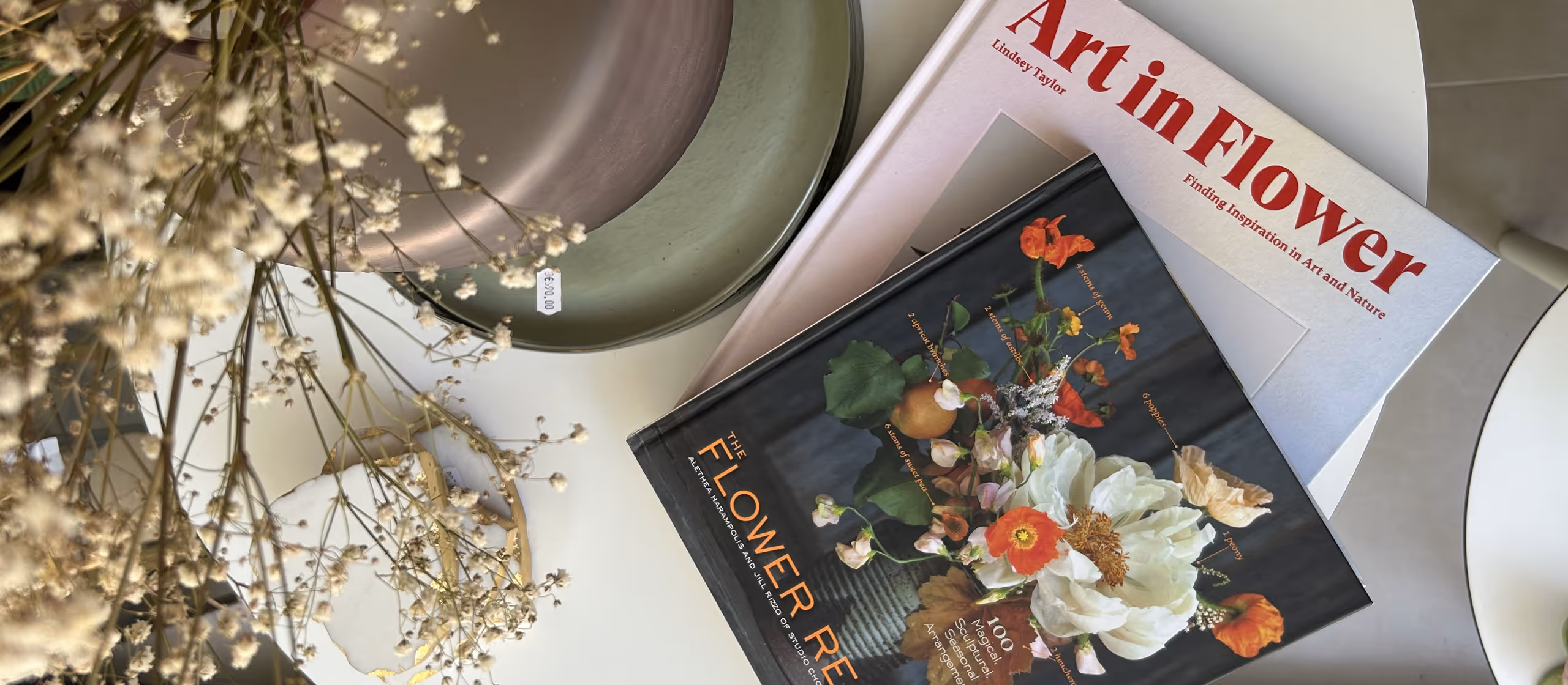 Two books on a white table with dried flowers and stacked ceramic plates in the background.