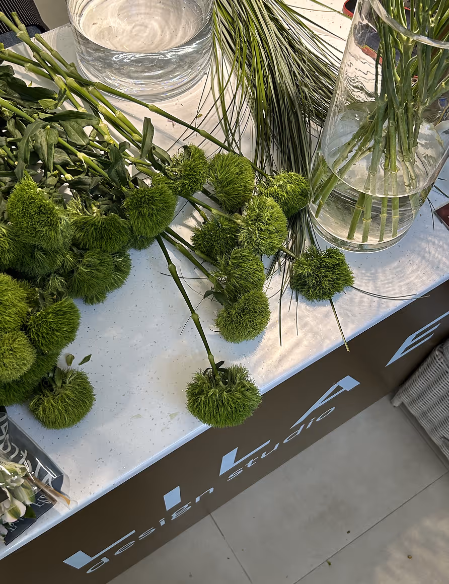 Green pom-pom flowers and long green leaves arranged on a white table next to a glass vase with flower stems.