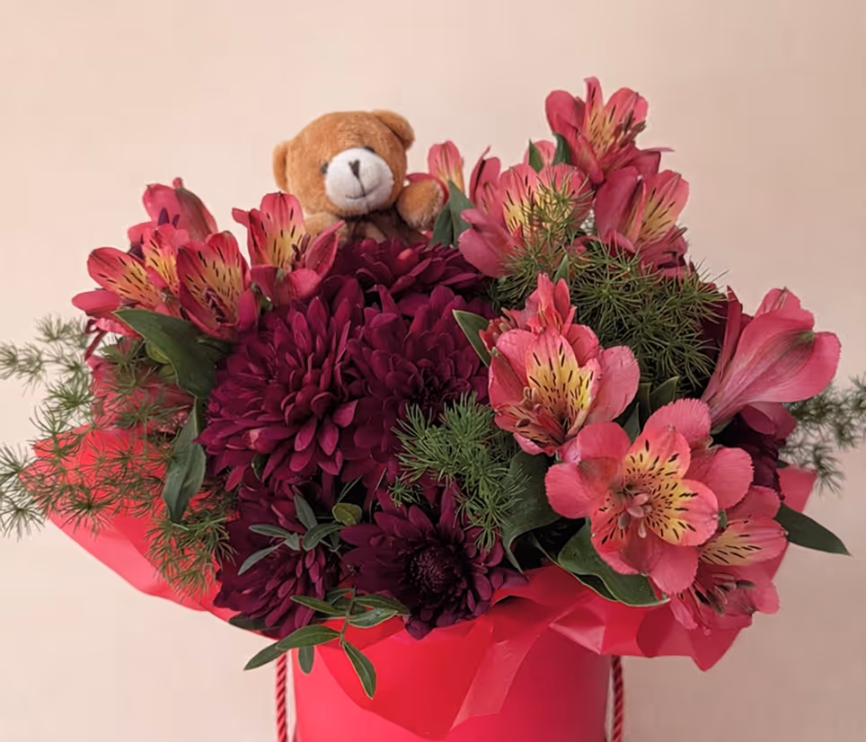 Red and purple flowers with green foliage in a red vase with a small teddy bear toy.
