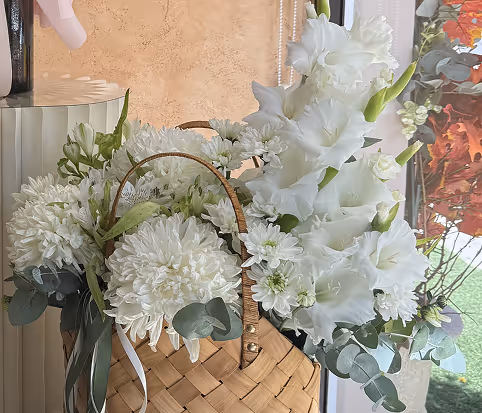Basket filled with white flowers including chrysanthemums and gladiolus with eucalyptus leaves, placed indoors by a window.
