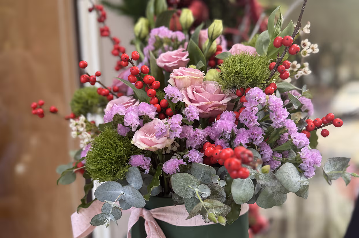 Bouquet of pink roses, purple flowers, red berries, green moss, and eucalyptus in a green vase with a pink ribbon.