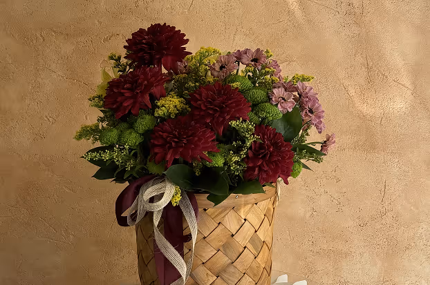 Bouquet of dark red and light purple flowers with green foliage in a woven basket decorated with maroon and white ribbons against a textured beige wall.