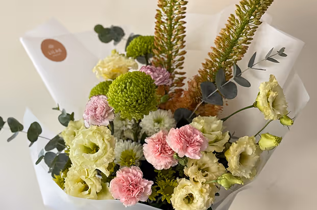 Bouquet of mixed flowers including pink carnations, green chrysanthemums, white lisianthus, and tall green foliage wrapped in white paper.