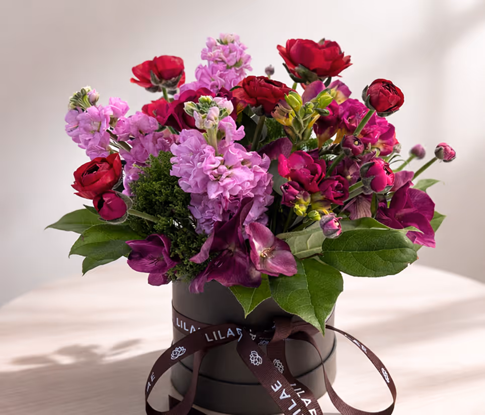 Bouquet of red, purple, and pink flowers with green leaves in a black round box with a ribbon.