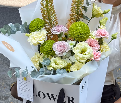 Bouquet of green chrysanthemums, white and pink carnations, and eucalyptus branches wrapped in white paper inside a white flower bag.