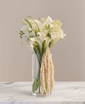 Clear vase with white calla lilies and hanging beige amaranthus on marble surface.