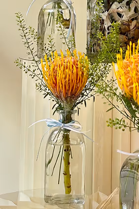 Orange protea flower with green leaves in a small clear glass vase tied with a blue ribbon.
