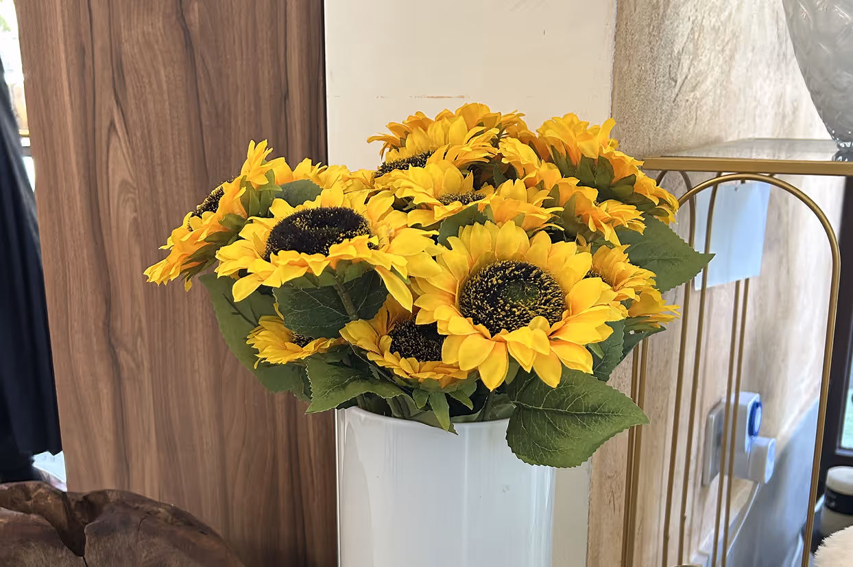 Bright yellow sunflowers with green leaves arranged in a white vase indoors near wood and glass surfaces.