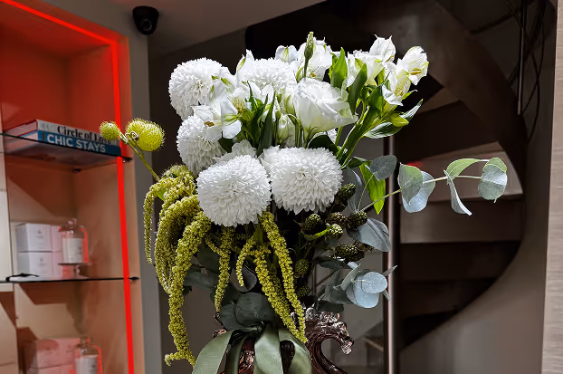 Elegant bouquet of white pom-pom flowers and green foliage arranged in a decorative vase with a dark background.