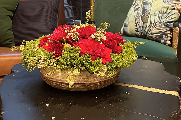 Decorative floral arrangement with red flowers and green foliage in a shallow round basket on a black coffee table.