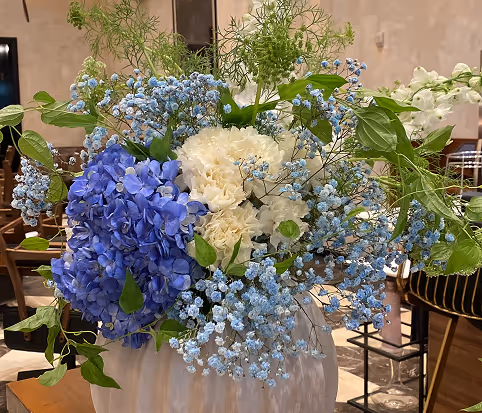 Floral arrangement with blue hydrangeas, white carnations, and small blue gypsophila flowers in a white vase on a wooden table indoors.