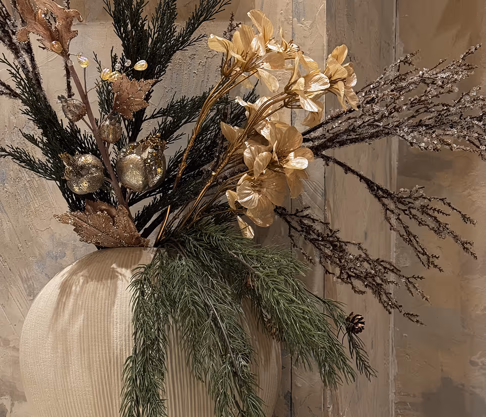Decorative vase holding green pine branches, golden flowers, and glittery autumn leaves against a textured wall.