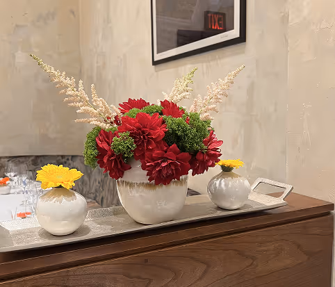 White ceramic pots on a wooden surface with red flowers and green foliage in the center pot, flanked by two smaller pots each with a single yellow flower.
