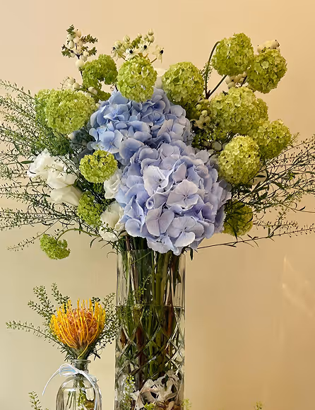Bouquet of blue hydrangeas and green viburnum flowers in a tall patterned glass vase.