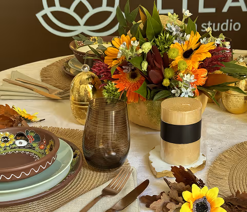 Elegant table setting with floral centerpiece featuring orange and yellow flowers, brown glass, decorative plates, and autumn leaves on woven placemats.