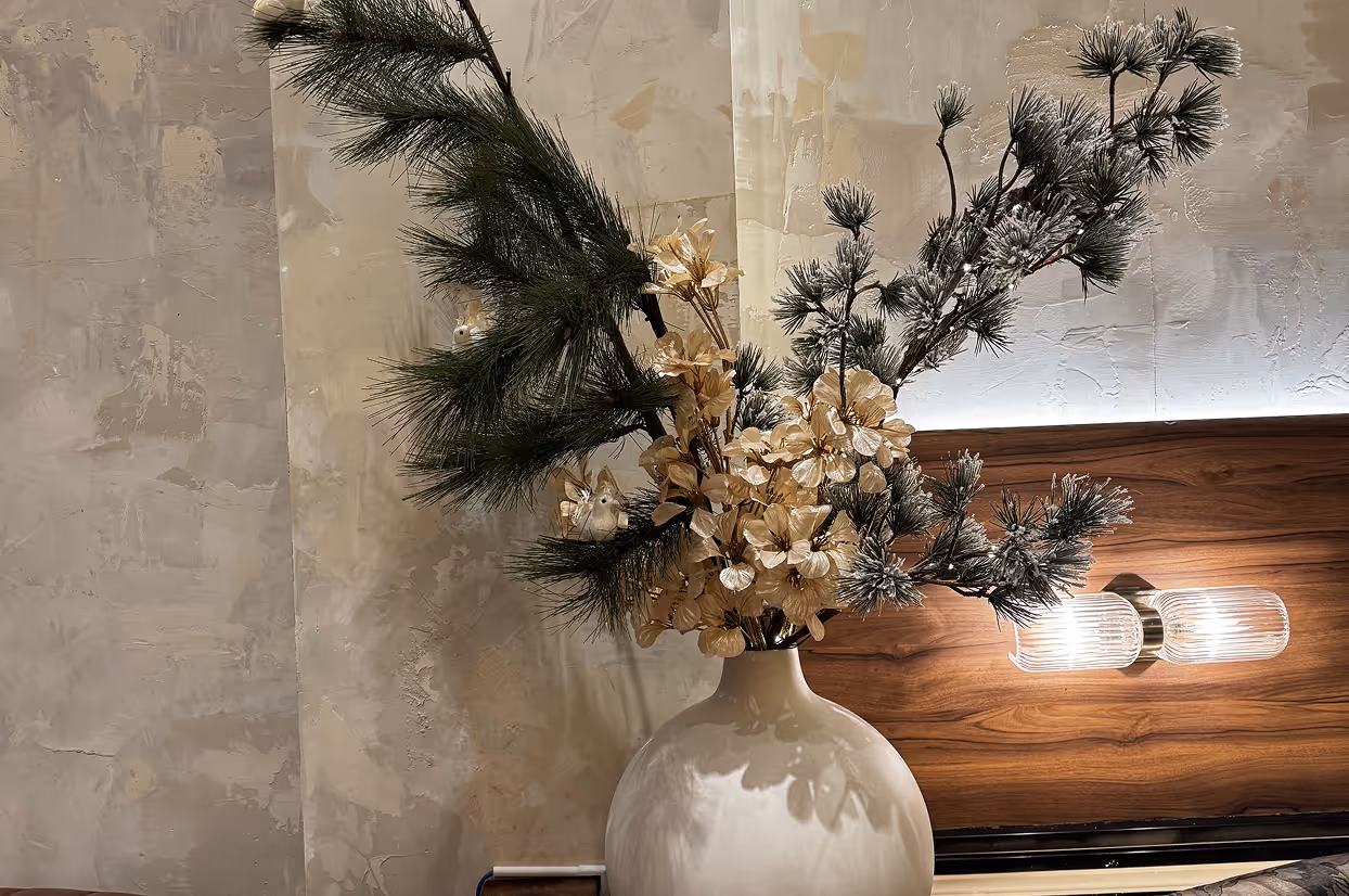 White vase holding pine branches and yellow flowers next to a lit bedside lamp on a wooden headboard.