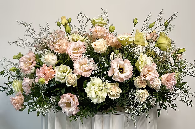Bouquet of pale pink and white lisianthus flowers with green foliage arranged in a white container against a plain background.